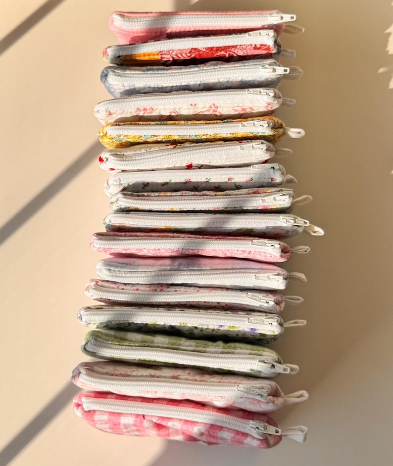 May include: A stack of small, rectangular fabric pouches with white zippers. The pouches feature various floral and patterned designs in shades of pink, red, yellow, blue, and green. The pouches are stacked vertically against a neutral background.