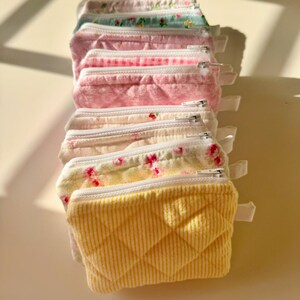 May include: A stack of eight small zippered pouches in various pastel colors and floral patterns. The pouches are made of quilted fabric and have a white zipper.