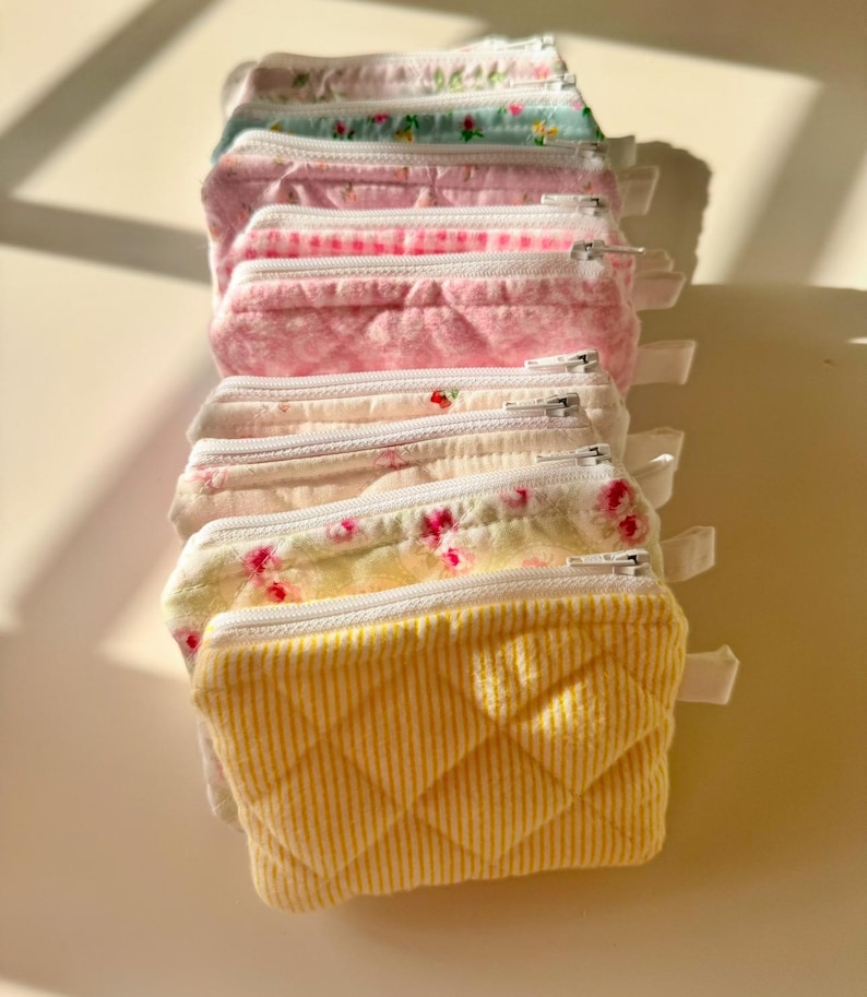 May include: A stack of eight small zippered pouches in various pastel colors and floral patterns. The pouches are made of quilted fabric and have a white zipper.