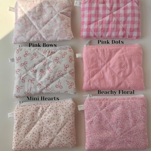 May include: A collection of small, quilted fabric pouches in various pink patterns. Designs include floral, gingham, polka dots, bows, and mini hearts. Each pouch has a zipper closure and a small fabric loop.