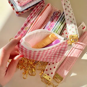 May include: A pink and white gingham fabric pouch with a zipper closure. The pouch is open and contains a small tube of hand cream and a small purple tube. The pouch is surrounded by a variety of pink and white fabric keychains with gold key rings.