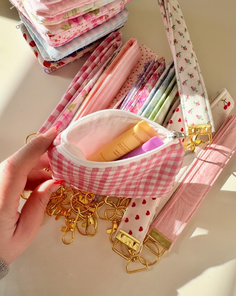 May include: A pink and white gingham fabric pouch with a zipper closure. The pouch is open and contains a small tube of hand cream and a small purple tube. The pouch is surrounded by a variety of pink and white fabric keychains with gold key rings.