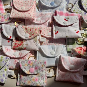 May include: A collection of small, fabric pouches with key rings. The pouches feature various floral, polka dot, and patterned designs in shades of pink, white, and blue. Each pouch has a button closure.