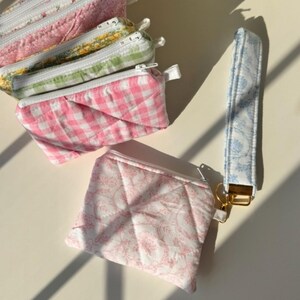 May include: A collection of quilted fabric pouches and a wristlet. The pouches feature floral, gingham, and patterned designs in pink, green, and white. The wristlet is light blue with a white floral pattern. The text "Mix & match with our wristlets!" is visible.