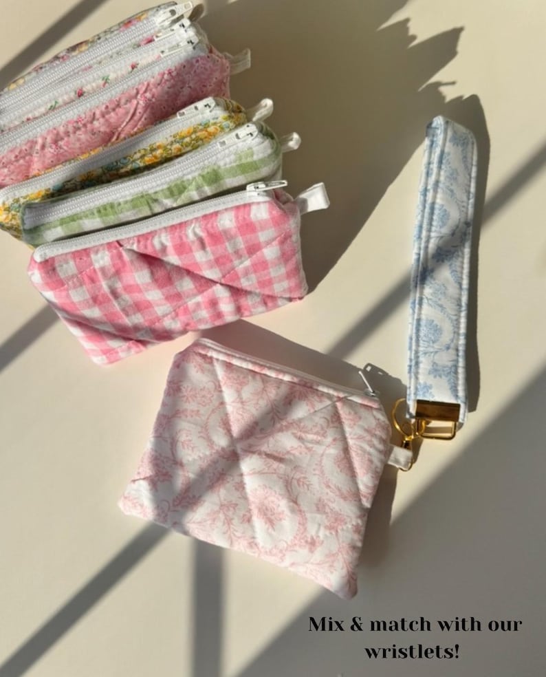 May include: A collection of quilted fabric pouches and a wristlet. The pouches feature floral, gingham, and patterned designs in pink, green, and white. The wristlet is light blue with a white floral pattern. The text "Mix & match with our wristlets!" is visible.
