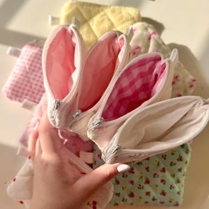 May include: Four small zippered pouches with different patterned fabric exteriors. The pouches are lined with white fabric and have white zippers. The pouches are stacked on top of each other.