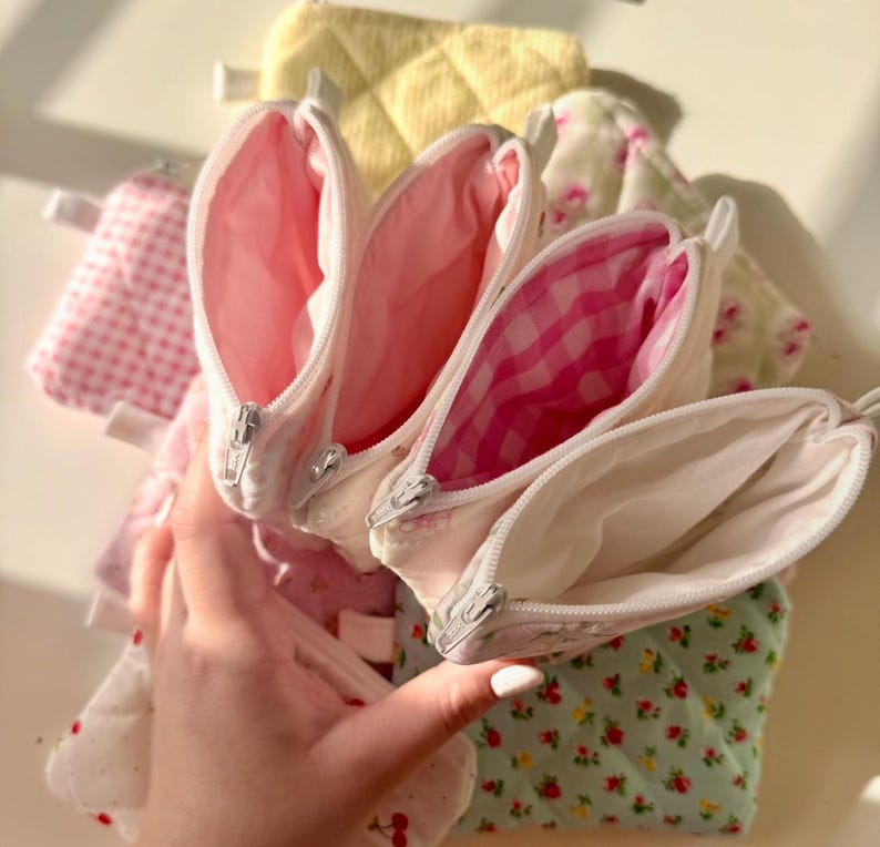 May include: Four small zippered pouches with different patterned fabric exteriors. The pouches are lined with white fabric and have white zippers. The pouches are stacked on top of each other.
