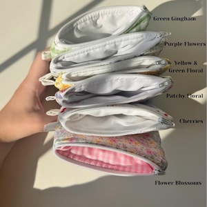May include: A stack of open, zippered pouches in various floral and patterned fabrics. The pouches include green gingham, purple flowers, yellow and green floral, patchy floral, cherries, and flower blossoms. Each pouch has a white interior.