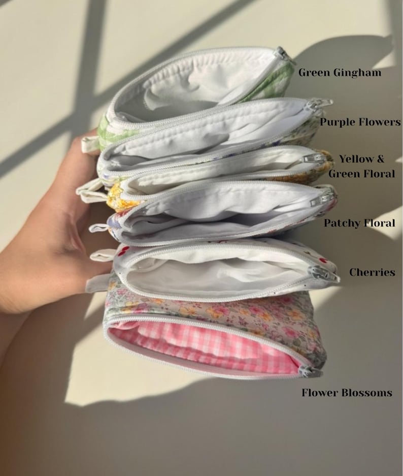 May include: A stack of open, zippered pouches in various floral and patterned fabrics. The pouches include green gingham, purple flowers, yellow and green floral, patchy floral, cherries, and flower blossoms. Each pouch has a white interior.