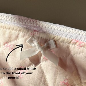 May include: A close-up of a white quilted fabric pouch with a white bow attached to the front. The pouch has a white zipper and a pink and white pattern of bows.