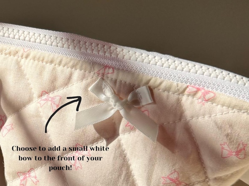 May include: A close-up of a white quilted fabric pouch with a white bow attached to the front. The pouch has a white zipper and a pink and white pattern of bows.