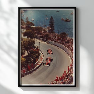 Può includere: Stampa incorniciata di una gara di Formula 1 a Monaco. Due auto da corsa rosse percorrono una curva, con una folla di spettatori. Lo sfondo presenta un mare blu e barche.