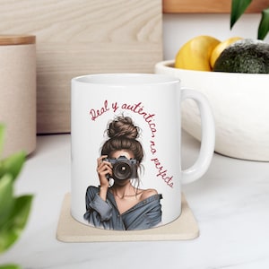 Könnte beinhalten: Weiße Keramik-Tasse mit einer Illustration einer Person, die eine Kamera hält, und dem Text "Real y auténtica, no perfecta" in roter Schrift. Die Person trägt ein Jeanshemd. Die Tasse hat einen weißen Griff.
