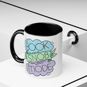 May include: White ceramic mug with a black interior and handle. The mug features the text "BOOK SNOB MODE" in a colorful design with blue, green, and purple accents. A perfect gift for book lovers.
