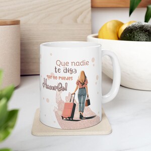 May include: White ceramic mug featuring a woman with blonde hair pulling a pink suitcase. The mug has the Spanish text "Que nadie te diga que no puedes hacer lo!" in black and pink. The mug is on a beige coaster.