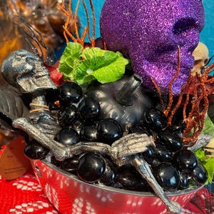 Può includere: Una statuetta di scheletro argentata si adagia in una ciotola argentata piena di perline di vetro nere. Un teschio di glitter viola si trova sopra una zucca nera, circondato da ramoscelli rossi e arancioni.