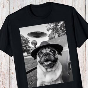 May include: Black t-shirt featuring a black and white image of a pug wearing a hat, with a UFO and an alien in the background. The pug is smiling. The image is printed on the front of the shirt.