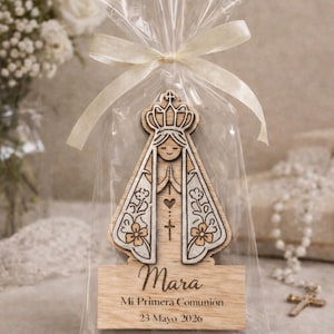 Può includere: Una statuetta religiosa in legno di una donna con una corona, in un sacchetto di cellophane trasparente legato con un nastro color crema. La statuetta ha un design a cuore e croce. Il nome "Mara" e il testo "Mi Primera Comunión 23 Mayo 2026" sono su una base di legno.
