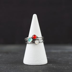May include: Stack of three silver rings with colorful gemstones: a red, a light blue, and a white stone. The rings are displayed on a white cone-shaped stand against a dark background.