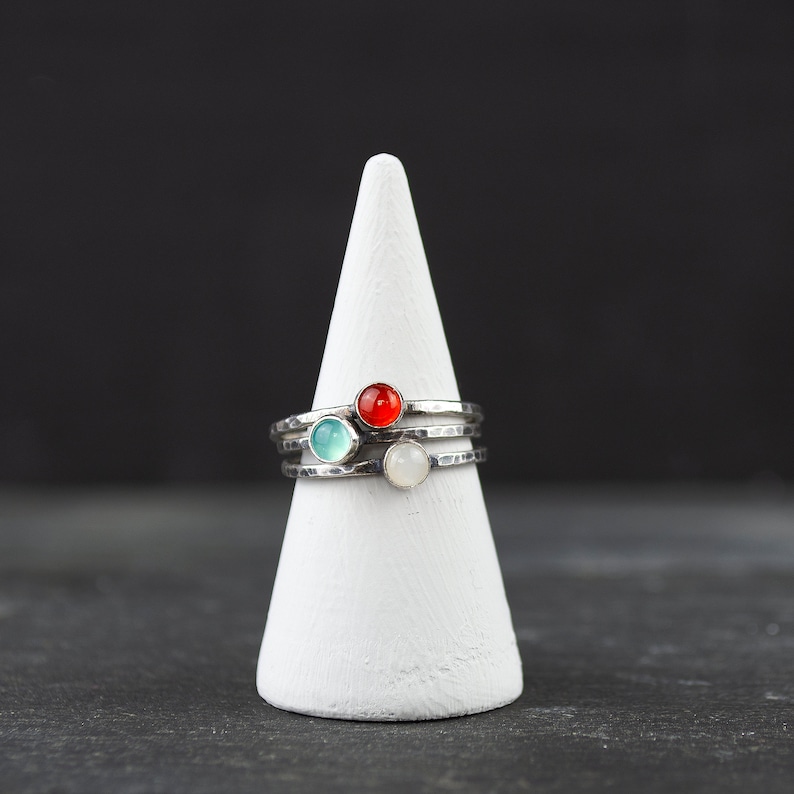 May include: Stack of three silver rings with colorful gemstones: a red, a light blue, and a white stone. The rings are displayed on a white cone-shaped stand against a dark background.