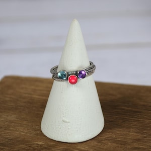 May include: A stack of three silver rings, each with a different colored gemstone: light blue, red, and purple. The rings are displayed on a white, cone-shaped jewelry holder, set against a wooden surface.