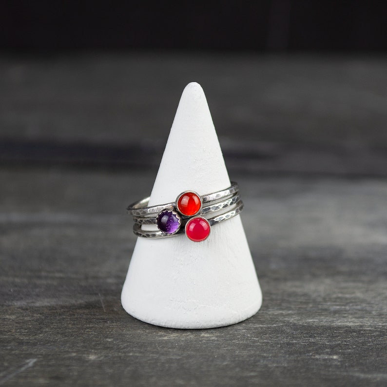 May include: A stack of three silver rings with hammered textures, each featuring a different colored gemstone: red, purple, and pink. The rings are displayed on a white, cone-shaped stand against a dark gray background.