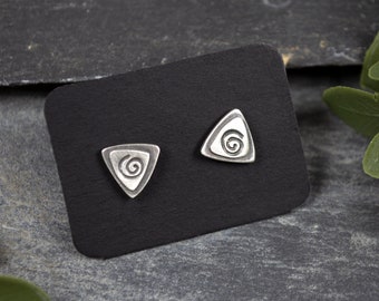 May include: A pair of silver-toned triangle stud earrings. Each earring features a raised spiral design within a triangular frame. The earrings are displayed on a black card against a dark gray stone surface.