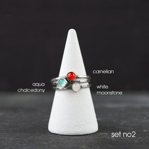 May include: A set of three sterling silver rings with hammered bands, each featuring a different gemstone: aqua chalcedony, carnelian, and white moonstone. The rings are displayed on a white cone-shaped stand against a dark background. Text on the image reads "set no.2".