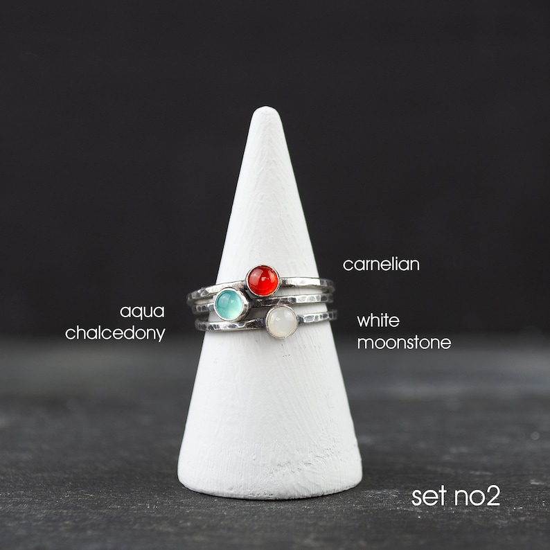 May include: A set of three sterling silver rings with hammered bands, each featuring a different gemstone: aqua chalcedony, carnelian, and white moonstone. The rings are displayed on a white cone-shaped stand against a dark background. Text on the image reads "set no.2".