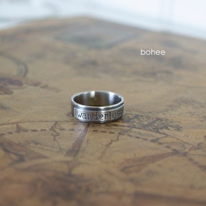 以下が含まれることがあります： 「wanderlust」の文字が刻まれたシルバーカラーのリング。リングはヴィンテージマップの背景に配置されています。ブランド名「bohee」が右上に見えます。