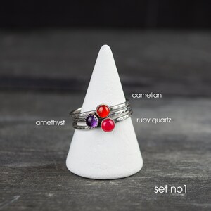 May include: Set of three silver rings with hammered bands, each featuring a different gemstone: amethyst, carnelian, and ruby quartz. The rings are displayed on a white cone-shaped stand against a dark background. Text on the image reads "carnelian", "ruby quartz", "amethyst", and "set no. 1".
