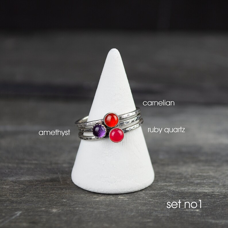 May include: Set of three silver rings with hammered bands, each featuring a different gemstone: amethyst, carnelian, and ruby quartz. The rings are displayed on a white cone-shaped stand against a dark background. Text on the image reads "carnelian", "ruby quartz", "amethyst", and "set no. 1".