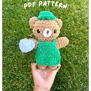May include: A tan crochet bear plushie wearing a green cap and a matching green apron. The bear has black eyes, a white muzzle, and holds a small white textured object. The text BEAR BARISTA PDF PATTERN is displayed in white above the plushie.