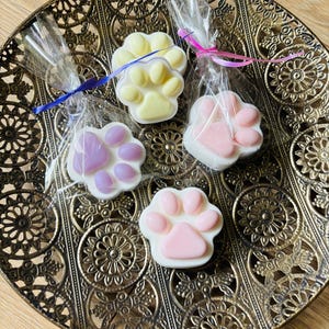 Op de afbeelding: Vier pootvormige zepen in pastelkleuren: geel, roze en lavendel. Elke zeep is verpakt in doorzichtig cellofaan en vastgebonden met een lint. De zepen zijn gerangschikt op een decoratief, bronskleurig dienblad.