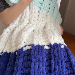 May include: A chunky knit blanket with three stripes of color: light blue, white, and dark blue. The blanket is made of soft, fluffy yarn and has a textured, cable knit pattern.