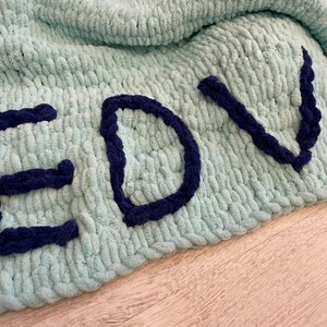 May include: A light blue, chunky knit blanket with the letters "EDV" in navy blue yarn. The blanket is made of a soft, plush material and is folded over. The letters are sewn onto the blanket.