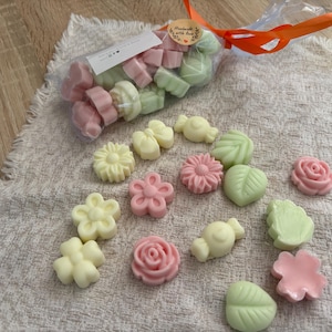 Op de afbeelding: Een verzameling pastelkleurige wassmeltstukken in bloemvorm. De smeltstukken hebben verschillende vormen, waaronder bloemen, bladeren en rozen. Sommige zitten in een transparante zak, vastgebonden met een oranje lint.