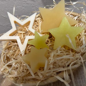 Può includere: Una collezione di saponi a forma di stella in un cestino di vimini pieno di paglia. I saponi variano di colore dal giallo pallido al crema. Una stella è un contorno vuoto. Il cestino è su una superficie di legno.