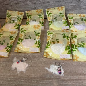 Könnte beinhalten: Mehrere kleine, transparente Plastiktüten gefüllt mit farbigen Pulvern, beschriftet mit Namen wie "Mango" und "Melody". Die Tüten haben ein Blumenmuster und sind auf einer Holzoberfläche angeordnet. Kleine Spielzeugautos und Schlüsselanhänger sind ebenfalls vorhanden.