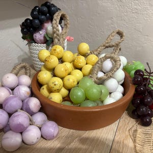 Può includere: Una varietà di uva artificiale in diversi colori, tra cui giallo, verde, bianco, viola e viola scuro. L'uva è disposta in una ciotola di terracotta e su una superficie di legno, con un piccolo cestino bianco di fiori sullo sfondo.