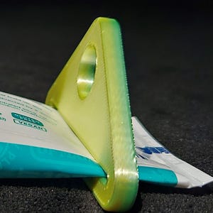 May include: A green and yellow 3D printed plastic tube squeezer. The squeezer is shaped like a triangle with a hole in the center. A tube of toothpaste is being squeezed through the hole.
