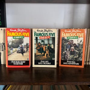 May include: Three vintage Enid Blyton 'Famous Five' books on a wooden shelf. The covers are orange and white, featuring illustrations and titles such as 'Five Get Into Trouble'. The books are displayed amongst other titles.