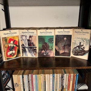 May include: A bookshelf displaying a collection of Agatha Christie mystery novels. The books are arranged in a row on the top shelf, with the titles "The Hollow", "The Murder of Roger Ackroyd", "Hickory Dickory Dock", "Lord Edgware Dies", and "Endless Night".