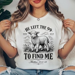 May include: White t-shirt with a black and white drawing of a lamb in a field. The text "HE LEFT THE 99 TO FIND ME" is above and below the lamb, with "Matthew 18:12-14" at the bottom. The shirt has a round neck.