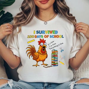 May include: White t-shirt with the text "I Survived 100 Days of School" in blue and yellow. A cartoon rooster with an open beak is next to a stack of books, scissors, and a pencil. The rooster says, "It's fine. I'm fine. Everything is fine."
