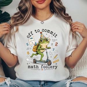 Könnte beinhalten: Ein weißes T-Shirt mit einer Grafik eines grünen Frosches, der einen Partyhut und eine Brille trägt und auf einem blauen Roller fährt. Der Frosch trägt einen gelben Rucksack. Der Text lautet off to commit math foolery, umgeben von Mathe-Symbolen.