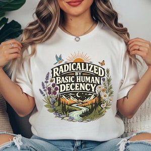 May include: White t-shirt with the text "RADICALIZED BY BASIC HUMAN DECENCY" in a circular design. The design includes a river, mountains, trees, flowers, a bird, and a butterfly. The shirt is made of a soft material.