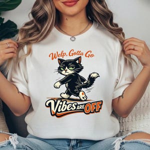 Puede incluir: Camiseta blanca con un gato de dibujos animados con expresión de enfado. El gato es negro y blanco con ojos verdes. El texto "Welp, Gotta Go" está encima del gato y "Vibes are OFF" debajo, en naranja y blanco.