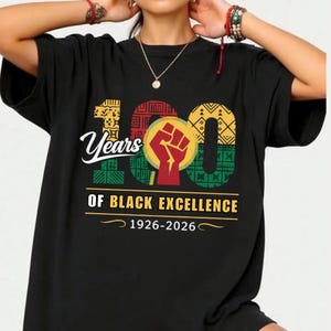 Könnte beinhalten: Schwarzes T-Shirt mit dem Text "100 Years of Black Excellence 1926-2026". Das Design beinhaltet eine geballte Faust, geometrische Muster und das Wort "Years" in einer Schreibschrift. Die Zahlen "100" sind in Gelb, Rot und Grün.