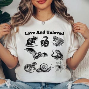 May include: A white t-shirt featuring a black graphic print of various animals including a rat, snake, frog, bat, lizard, gecko, snail, and spider web. The text "Love And Unloved" is printed in black above the collection of animal illustrations.